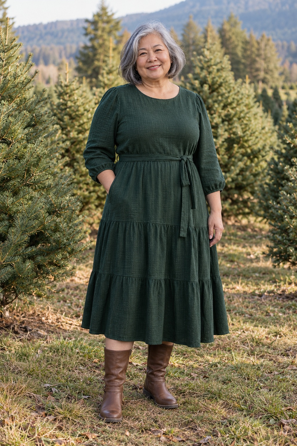 A gray-haired woman wears a forest-green dress and brown boots at a tree farm.